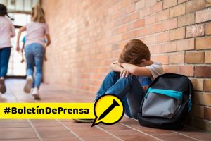 Little boy sitting alone on floor after suffering an act of bullying while children run in the background. Sad young schoolboy sitting on corridor with hands on knees and head between his legs.